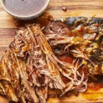 Slow cooker brisket partly pulled on a wooden board with soft onions and a bowl of cooking juices