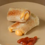 Two grilled cheese roll-ups stacked on a beige plate, with melted orange cheese at their open ends and red dip beside them