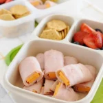 Lunchbox ham and cheese rolls with visible cheddar ends, packed beside crackers, berries, and corn