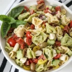 White bowl of cold green and yellow tortellini salad with tomatoes, mozzarella, salami, olives, and basil