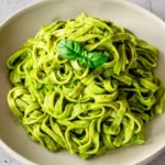 Spinach fettuccine coated in smooth green sauce, mounded in a shallow cream bowl with basil on top