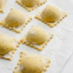 Uncooked cheese and spinach ravioli with rounded centers and fluted square edges on lightly floured parchment