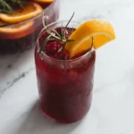 Tall glass of ruby cranberry punch with ice, cranberries, orange, and rosemary; a pitcher sits behind it
