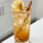 Caramel apple mocktail with ice, caramel streaks, a sugared rim, apple slices, and a cinnamon stick