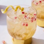 Pale golden sparkling apple cider mocktail over ice with pomegranate arils and a curled lemon peel