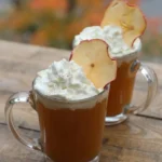 Two clear mugs of hot spiced apple cider topped with whipped cream and upright dried apple slices