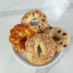 Four mini protein bagels on a white plate with everything seasoning, cheddar, sprinkles, and raisins