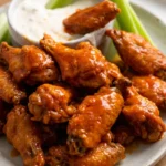 Glossy Buffalo chicken wings piled on a light platter with celery and blue cheese dressing