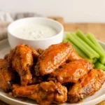 Glossy Buffalo chicken wings piled on a light platter with celery and blue cheese dressing