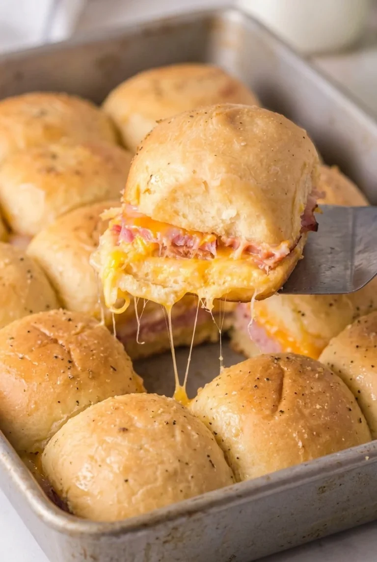 Ham and Cheese Hawaiian Roll Sliders A golden ham and cheese Hawaiian roll slider lifted above a pan, with pink ham and melted cheese showing.