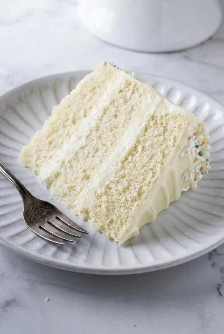 A generous slice of sugar-free vanilla cake with ivory cream cheese frosting and colorful sprinkles on a ridged plate
