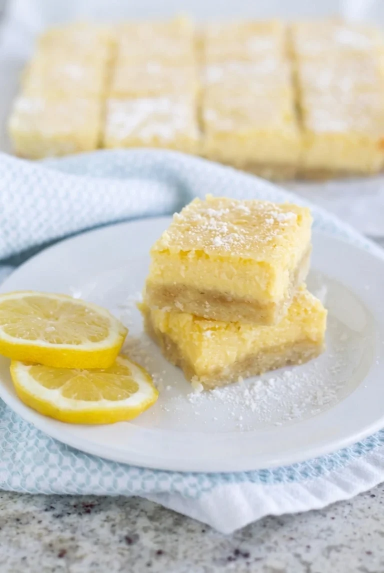 Sugar-Free Lemon Bars with Almond Flour Sugar-free lemon bars stacked on a white plate with a pale crust, yellow filling, powdered sweetener, and lemon slices