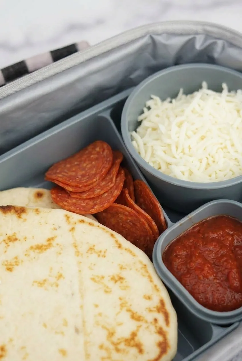 Close view of browned mini flatbread, pepperoni, shredded mozzarella, and pizza sauce in a lunchbox