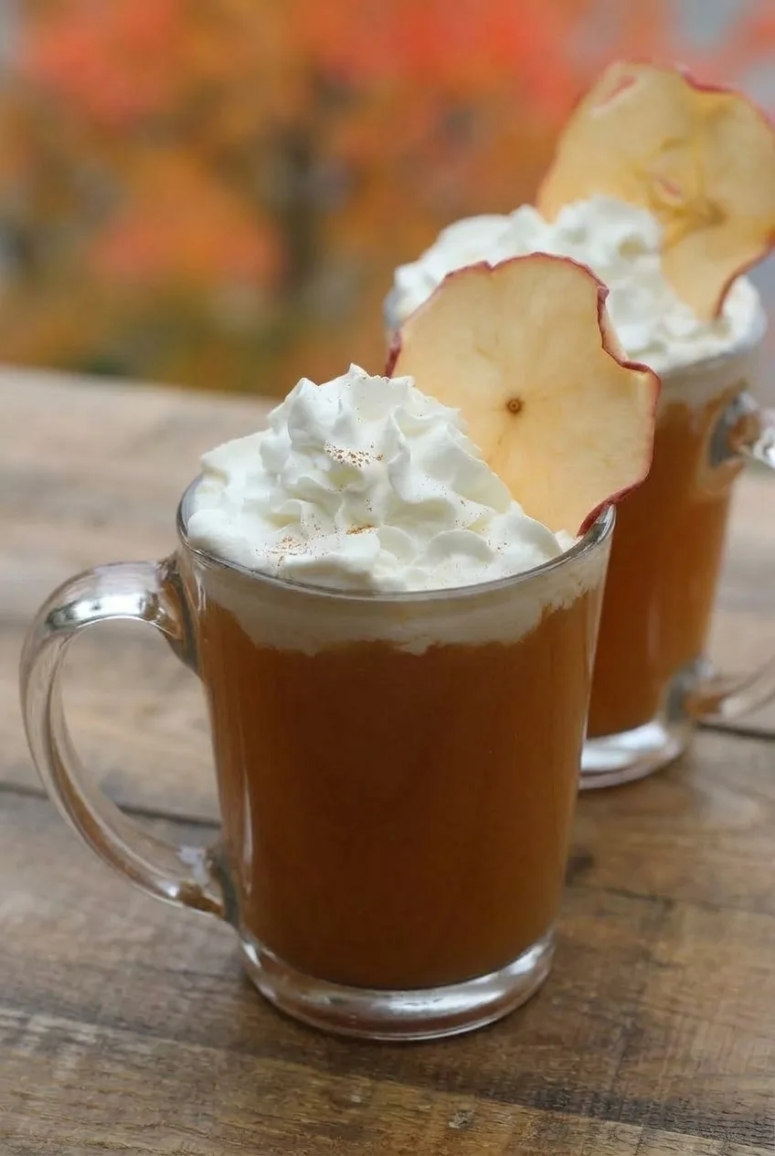 Close view of amber spiced cider beneath whipped cream and an upright red-edged apple slice