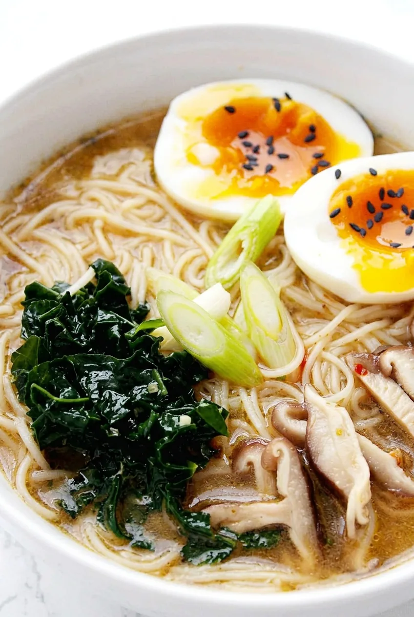 Close view of ramen noodles, sliced shiitake mushrooms, wilted kale, and sesame-topped jammy egg halves
