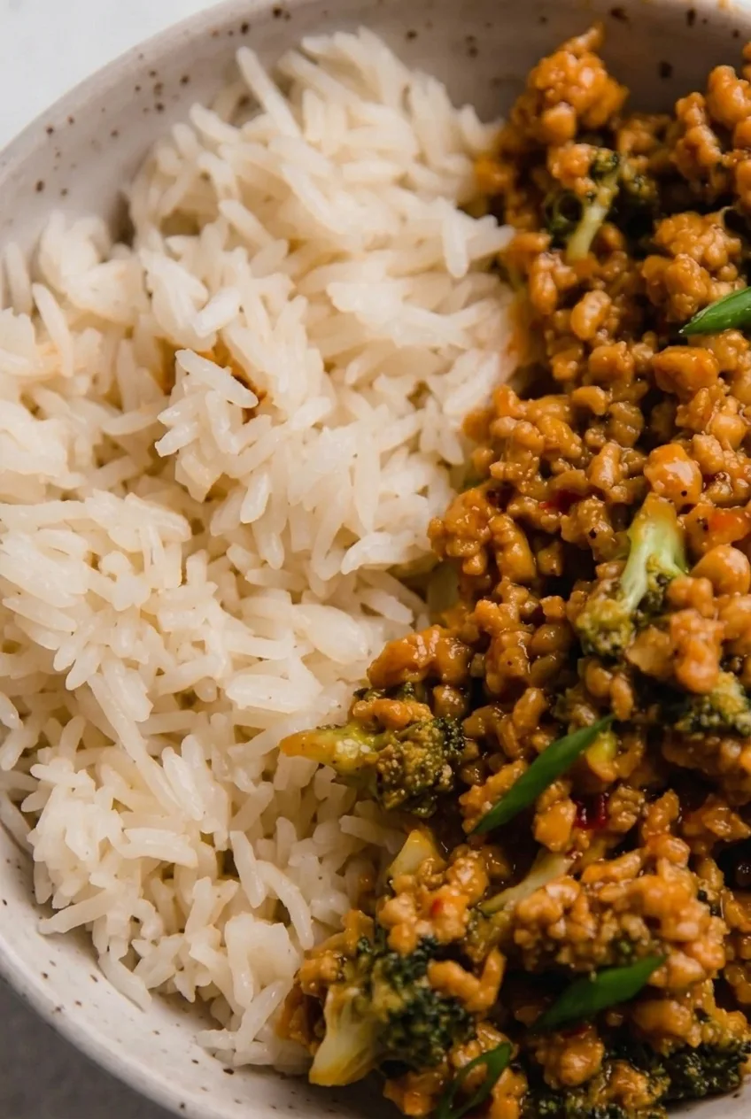 Close view of glossy ground chicken crumbles, broccoli florets, green onion slices, and distinct rice grains