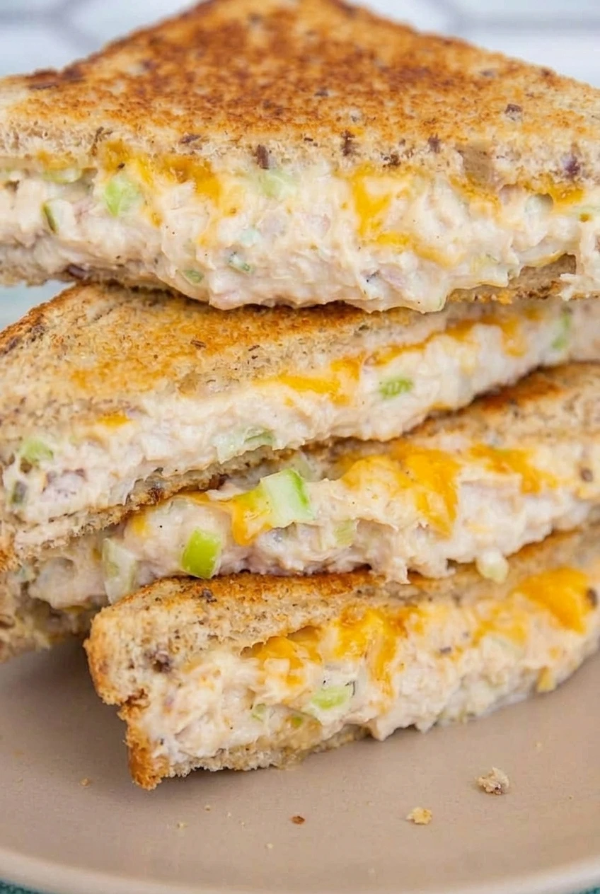 Close-up of a tuna melt cut edge showing creamy tuna, celery flecks, melted cheddar, and toasted multigrain bread.