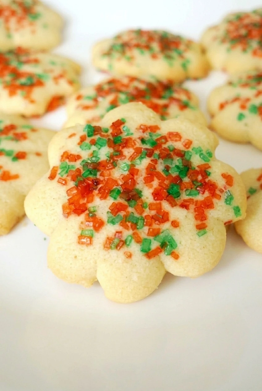 Close detail of a pale almond spritz cookie showing rounded petals, a lightly golden edge, and red and green sprinkles