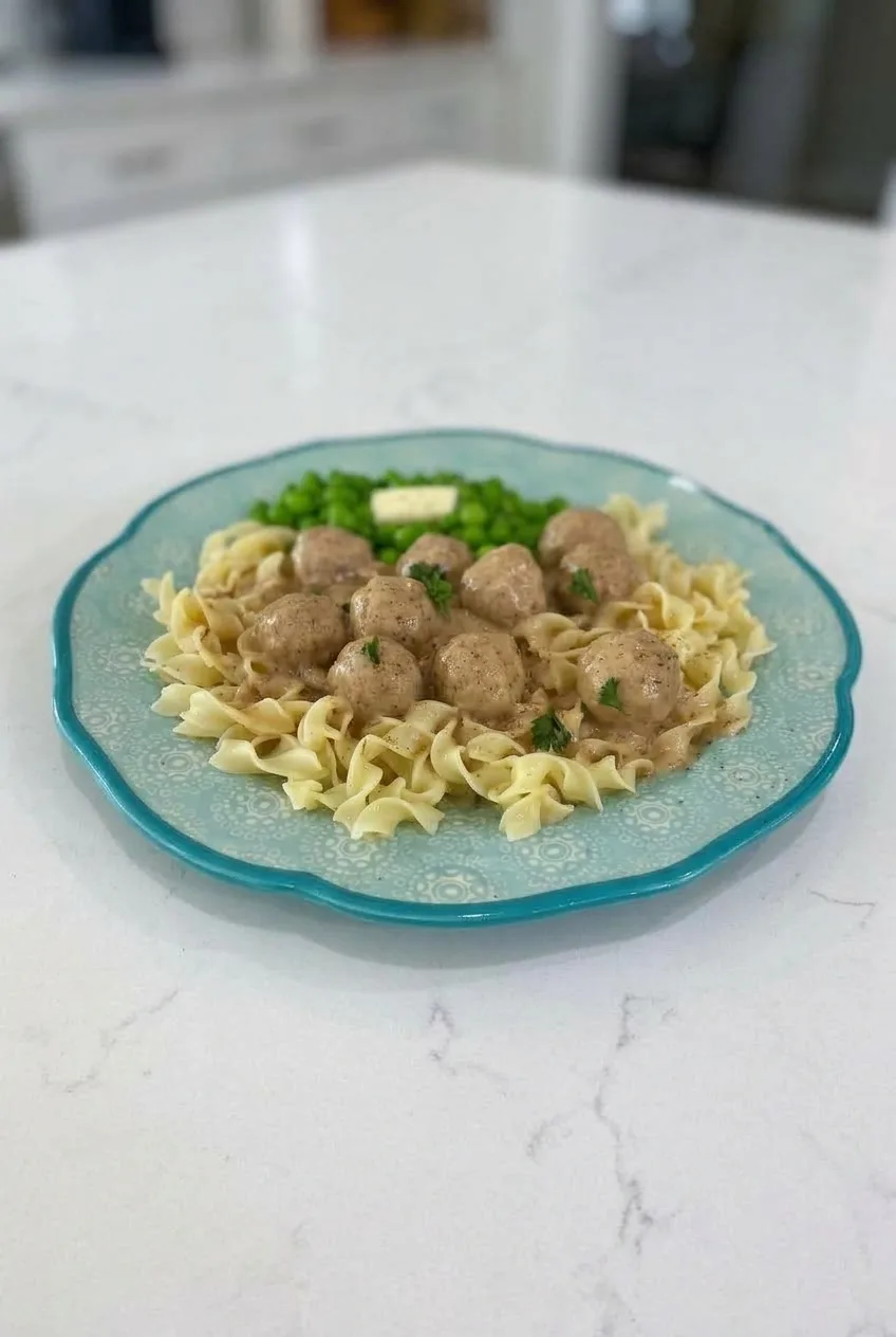 Full plate of Swedish meatballs over egg noodles with a separate mound of peas on a light countertop