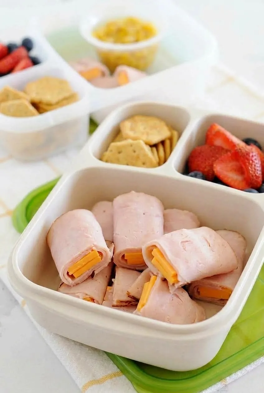 Divided lunchbox of ham and cheese rolls with crackers, berries, and corn on a light surface