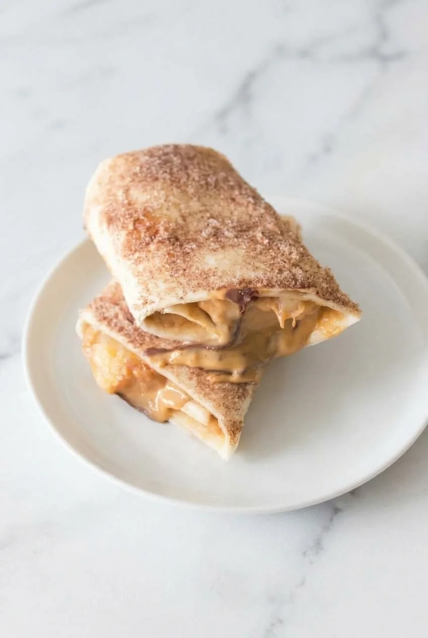 Two stacked cinnamon-sugar banana roll-up halves on a pale plate, seen in a wider view