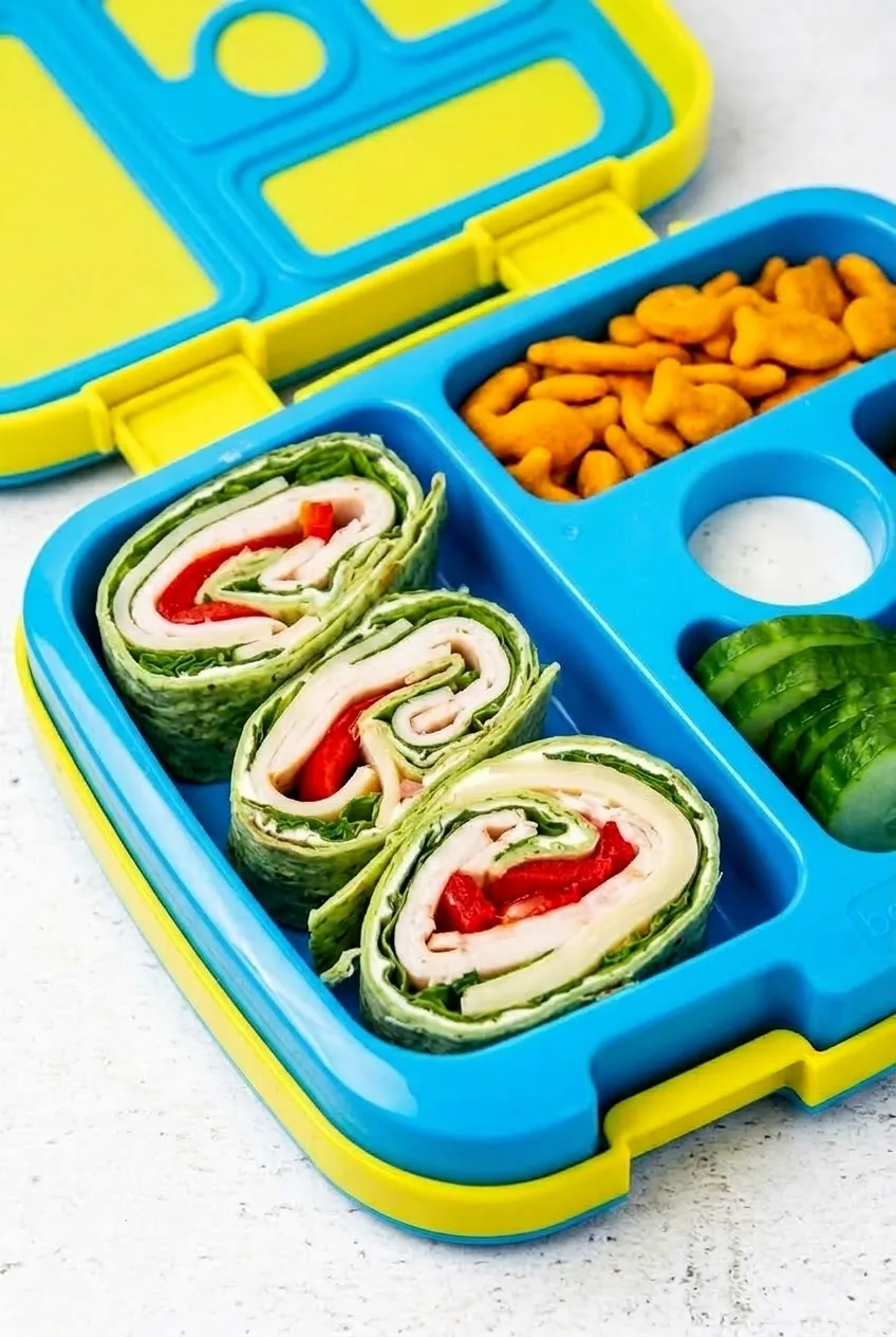 Open divided lunchbox with turkey and cheese pinwheels, cucumber, crackers, and dip