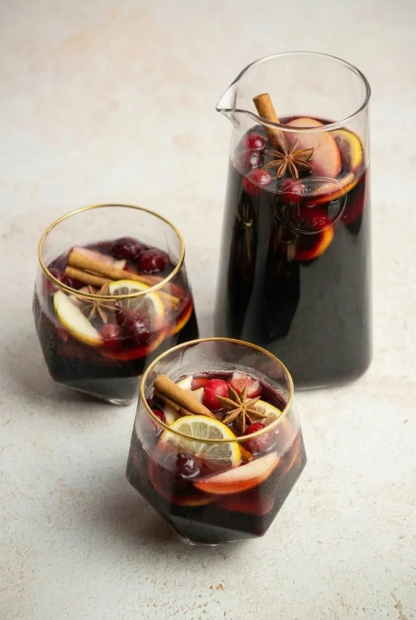 Pitcher and two garnished glasses of deep-red Thanksgiving sangria on a light tabletop