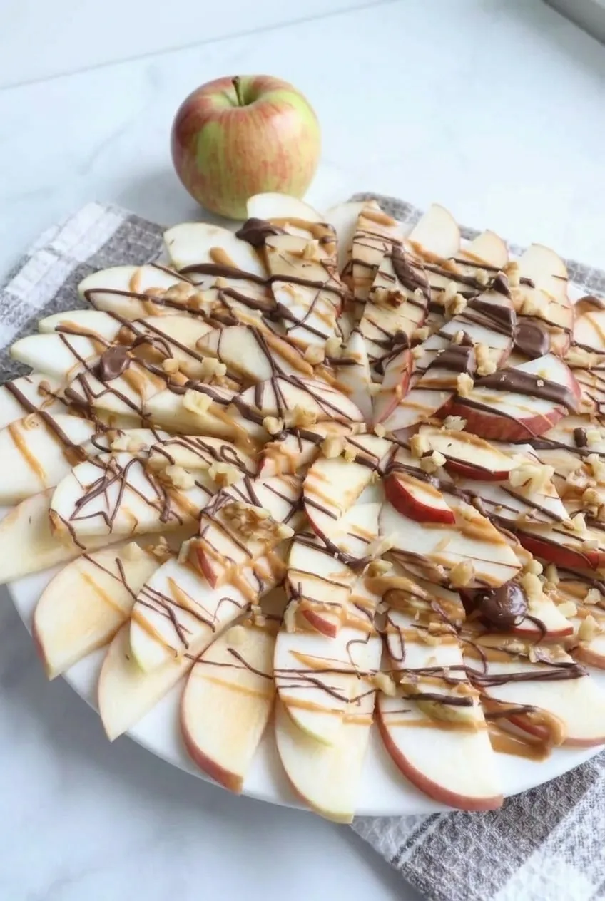 Full platter of apple slices with peanut butter, chocolate, and walnuts on a light surface