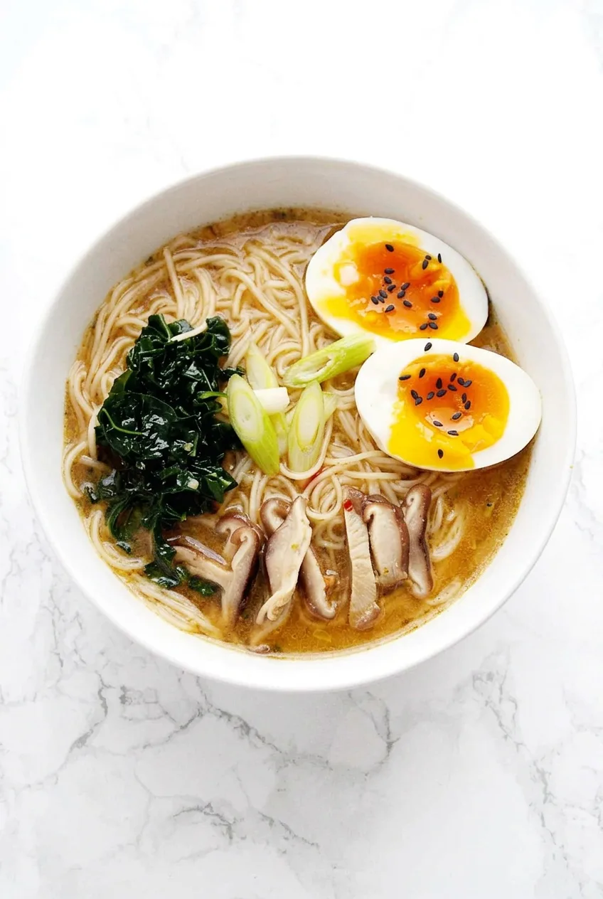 Complete bowl of vegetable ramen with noodles, kale, mushrooms, scallions, and jammy egg on pale marble