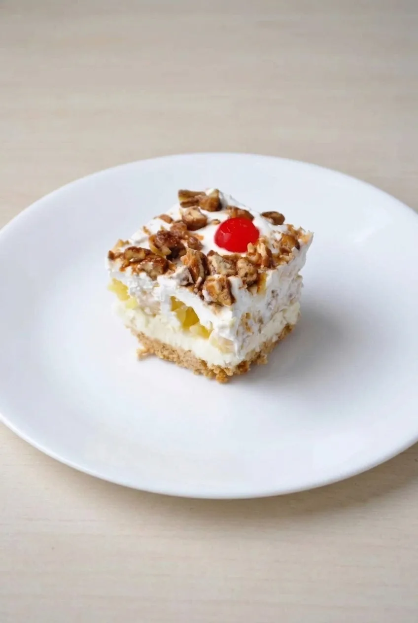 A plated square of banana split dessert with pecans and a cherry, shown with more of the plate and tabletop