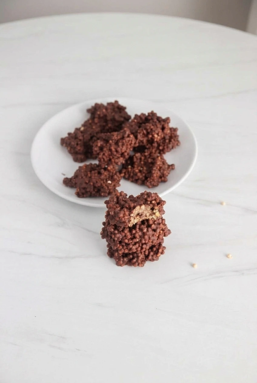 A full serving of irregular dark chocolate quinoa crunch bites on a white plate, including one split-open cluster.