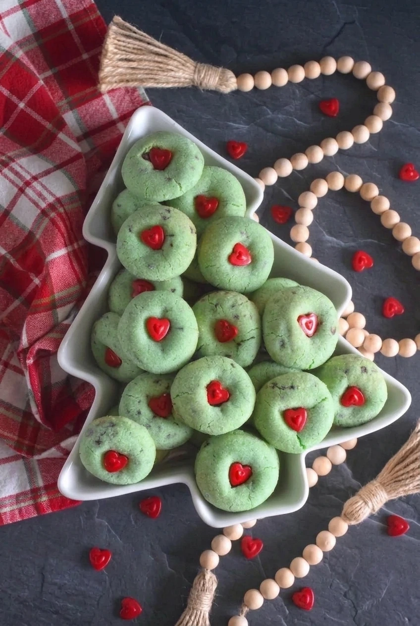 Full platter of round green Grinch cookies with red hearts on a dark slate surface