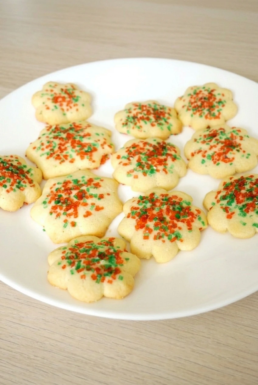 A full white plate of flower-shaped almond spritz Christmas cookies with red and green sprinkles